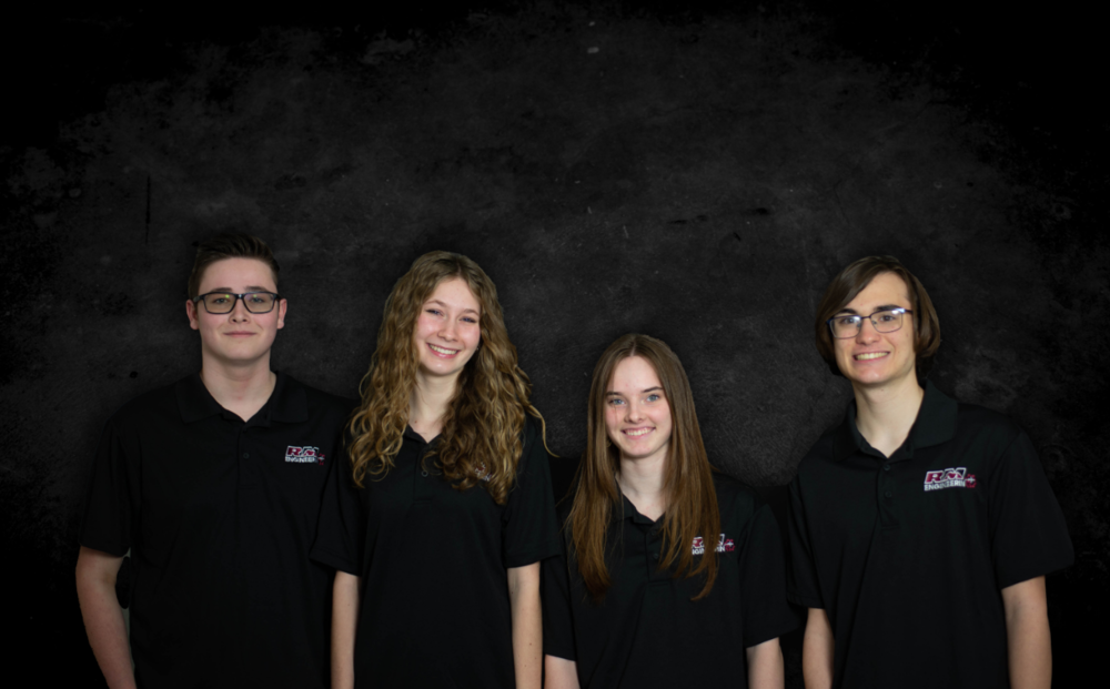 Photograph of four students against a black textured background, lined up next to each other and cropped at the waist. All students are wearing black RM Engineering polos.