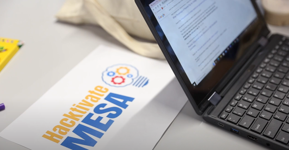 Image of an open laptop with Google results on the screen, tabletop with Hacktivate Mesa logo printed on a piece of paper, the corner of a coloring pencil box, and part of a bag in the background